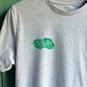 Może przedstawiać: Szary t-shirt z zielonym nadrukiem dwóch plasterków ogórka.