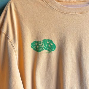 Może przedstawiać: Jasnożółty t-shirt z długim rękawem z zielonym nadrukiem dwóch plastrów ogórka.