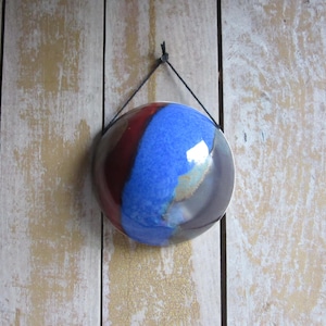 May include: A circular ceramic wall ornament featuring a glossy glaze in shades of deep blue, dark red, and grey. The piece is suspended by a thin black cord attached to a small loop at the top, creating a smooth, reflective surface finish.