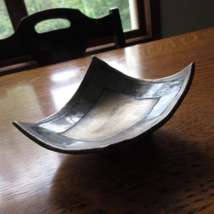May include: A square ceramic dish with a black glaze and a silver metallic sheen. The dish is slightly curved and has a unique shape.