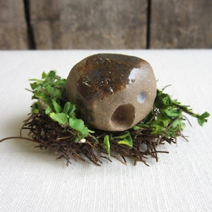 May include: A small, brown ceramic house with a hole in the front, nestled in a bed of brown twigs and green leaves.