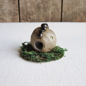 May include: A small, round, gray ceramic sculpture with a hole in the side and black beads on top. The sculpture is sitting on a bed of green moss.