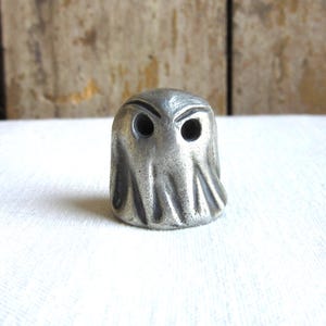 May include: A small, silver, pewter figurine of a ghost with two eye holes. The ghost is standing on a white surface.