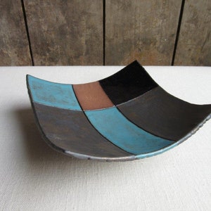May include: A square ceramic dish with a geometric pattern of black, brown, and blue glaze. The dish is slightly curved and has a rustic, handmade look.