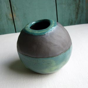 May include: A small, round ceramic vase with a gray, teal, and white glaze. The vase has a wide opening at the top and a rounded bottom.