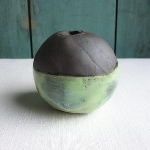 May include: A small, round ceramic vase with a dark gray top and a light green bottom. The vase has a smooth, glossy finish.