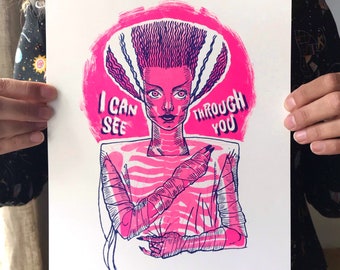 Puede incluir: Una impresión artística rosa y morada que presenta una ilustración estilizada de la Novia de Frankenstein. La obra de arte incluye el texto "I CAN SEE THROUGH YOU" y está ambientada sobre un fondo circular rosa. El pelo de la novia es morado.