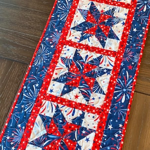 May include: A patriotic table runner featuring a repeating pattern of red, white, and blue stars and fireworks. The central design showcases large star shapes, framed by a red border and a blue background with star and firework motifs.