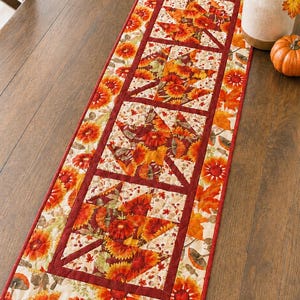 May include: A rectangular table runner with a fall theme. The runner features a repeating pattern of orange and red flowers, leaves, and mushrooms on a cream background, framed by a dark red border. The runner is approximately 152 cm long.