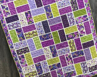May include: A purple and green patchwork quilt with a geometric pattern. The quilt is made of various fabrics with floral prints and different textures.