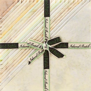 May include: A stack of 20 cream-colored fabric squares with a green and black ribbon tied around them. The ribbon has the text "Island Batik" printed on it.