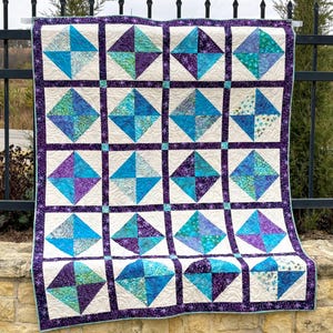 May include: A handmade quilt with a geometric diamond pattern. The quilt features squares of white fabric with alternating turquoise and purple diamond shapes. The quilt is bordered with a purple fabric with a star pattern.