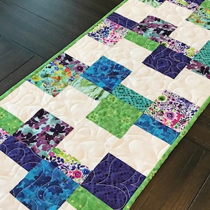 May include: A green, white, and purple patchwork table runner with a quilted design. The runner is made of fabric squares with floral patterns.