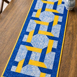 May include: A rectangular table runner with a geometric design. The runner features a blue and yellow pattern on a light blue floral background. The edges are bordered with a solid blue fabric and a yellow trim. The runner is approximately 12 inches wide and 36 inches long.