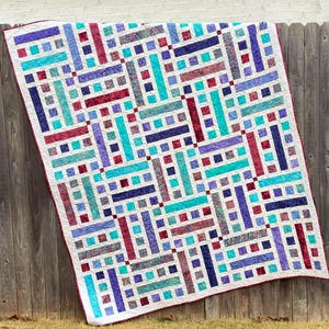 May include: A handmade quilt with a geometric pattern in shades of teal, purple, burgundy, and navy blue. The quilt features a white background with a repeating design of rectangles and squares. The edges are finished with a red binding.