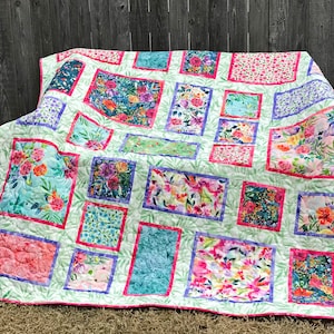 Fancy Frames Quilt pattern of watercolor floral squares and rectangles framed with pink and purple fabric on a light green background. Quilt is shown folded on a bench.