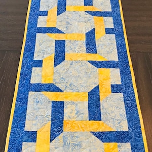 May include: A blue and yellow table runner with a geometric pattern. The runner is made of fabric and has a quilted texture.