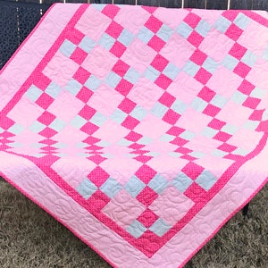 Op de afbeelding: Een roze en witte patchworkdeken met een ruitpatroon. De deken is gemaakt van verschillende stoffen, waaronder een roze stof met witte stippen en een witte stof met een subtiel patroon.