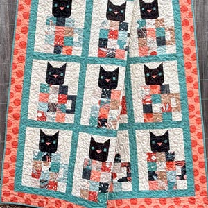 Pode incluir: Uma colcha com um design de patchwork que apresenta gatos pretos em cada quadrado. A colcha tem uma borda turquesa e uma borda coral e branca com bolinhas.