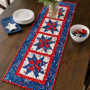 May include: A patriotic table runner with a star and firework design in red, white, and blue. The runner features a central panel with large star patterns, framed by red borders and a blue background with firework motifs. It is placed on a dark wooden table.
