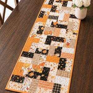 May include: A rectangular table runner with a patchwork design. The runner features a mix of orange, black, and cream-colored fabrics with pumpkin, plaid, and floral patterns. The edges are trimmed with a solid orange border. A vase of white flowers sits on the table.