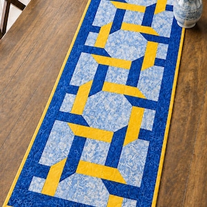 May include: A rectangular table runner with a geometric design. The runner features a blue and yellow pattern with a floral print background. The border is a solid blue with a yellow edge. The runner is approximately 30 cm wide and 91 cm long.
