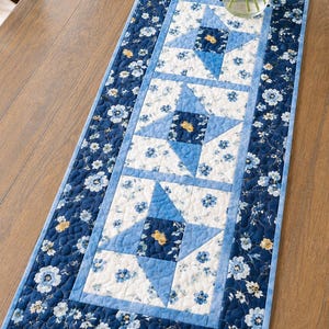 Pode incluir: Caminho de mesa floral azul e branco com um padrão de estrelas no centro. O caminho de mesa é feito de tecido acolchoado e tem uma borda floral. O caminho de mesa tem aproximadamente 30 cm de largura e 91 cm de comprimento.