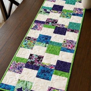 Può includere: Runner da tavolo trapuntato rettangolare con un design patchwork. Il runner presenta file alternate di quadrati bianchi e quadrati con motivi floreali e geometrici nei toni del blu, verde e viola. Un bordo verde incornicia il runner.