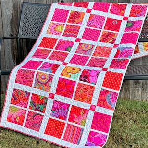 May include: A pink and white quilt with a floral pattern. The quilt is made up of squares with different floral designs. The quilt is sitting on a wooden bench.