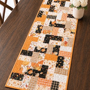 May include: A rectangular table runner with a patchwork design. The runner features a mix of orange, black, and cream-colored fabrics with pumpkin, plaid, and floral patterns. The edges are trimmed with a solid orange border. A vase of white flowers sits on the table.