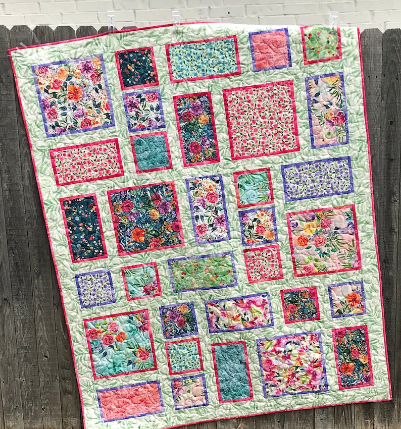 Fancy Frames Quilt pattern of watercolor floral squares and rectangles framed with pink and purple fabric on a light green background. Quilt is shown hanging on a fence.