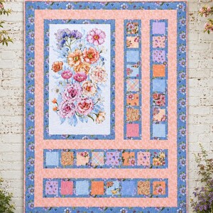 May include: A rectangular quilt featuring a large central panel of watercolour-style pink, orange, and purple flowers on a white background, surrounded by peach-coloured fabric borders and rows of small square patches in floral and plaid patterns.