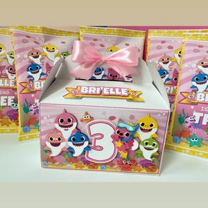 May include: Pink and white party favor box decorated with cartoon sharks, the number 3, and the name "Bri'Elle". The box is tied with a pink ribbon and surrounded by matching favor bags.