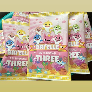 May include: Multiple rectangular snack bags with a light pink and yellow striped background featuring cartoon sharks, colorful coral, and sea life. The text on the bags reads BRI'ELLE IS TURNING THREE in white and yellow lettering.