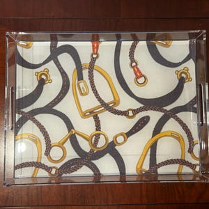 May include: A rectangular clear acrylic serving tray featuring a white base with a repeating pattern of dark gray straps, braided leather cords, and gold-toned equestrian hardware including stirrups and bits, with integrated side handles.