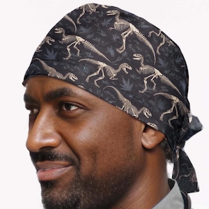 May include: Black surgical cap with a repeating pattern of dinosaur skeletons in tan against a dark background. The cap is tied at the back and is worn by a person wearing a gray scrub top.