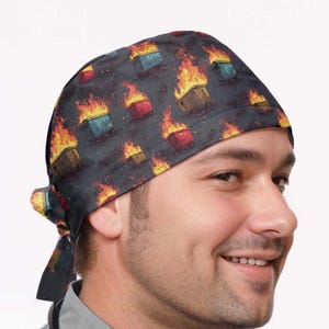May include: A dark gray surgical scrub cap featuring a repeating pattern of burning dumpsters in shades of red, yellow, blue, and brown. The cap is tied at the back.