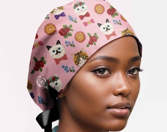 May include: A pink scrub cap featuring a repeating pattern of illustrated cats wearing bow ties, floral wreaths, and interspersed with red roses and golden donuts. The cap includes a clear side button and a black ribbon tie at the back for adjustment.