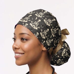 May include: A black scrub cap featuring a repeating pattern of off-white skeletons in various yoga poses, including lotus position. The cap has a gathered back design with a solid tan ribbon tie closure, providing a secure and adjustable fit.