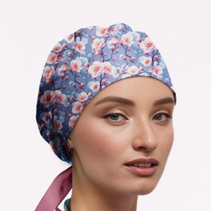 May include: A scrub cap featuring a blue background with a repeating pattern of pink and white cherry blossom flowers and dark branches. The cap includes a solid pink ribbon tie at the back, designed to secure the fit around the head comfortably.