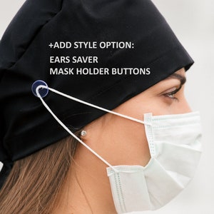 K&ouml;nnte beinhalten: Eine schwarze OP-M&uuml;tze mit wei&szlig;en Gummib&auml;ndern und einem durchsichtigen Knopf an der Seite. Auf der M&uuml;tze steht der Text "ADD STYLE OPTION: EARS SAVER MASK HOLDER BUTTONS". Eine Person tr&auml;gt die M&uuml;tze und eine hellblaue OP-Maske.