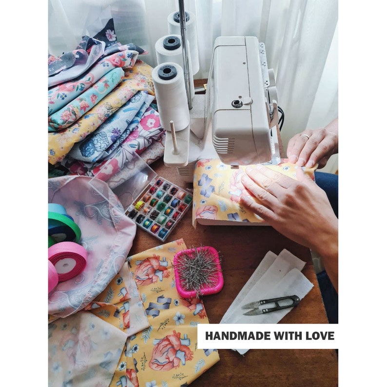 K&ouml;nnte beinhalten: Nahaufnahme einer N&auml;hmaschine mit Stoff und Garnrollen. Der Stoff ist gelb mit einem Blumenmuster und der Text "HANDMADE WITH LOVE" ist sichtbar.