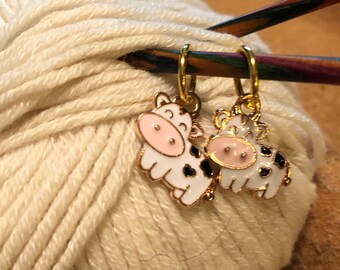 May include: Two gold cow stitch markers with black and white spots and pink accents. The markers are attached to a wooden knitting needle.
