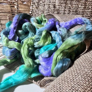 May include: A pile of colorful fiber in shades of teal, green, and purple. The fiber is arranged on a burlap-like fabric, with a woven basket in the background. The fiber appears soft and fluffy, suitable for crafting.