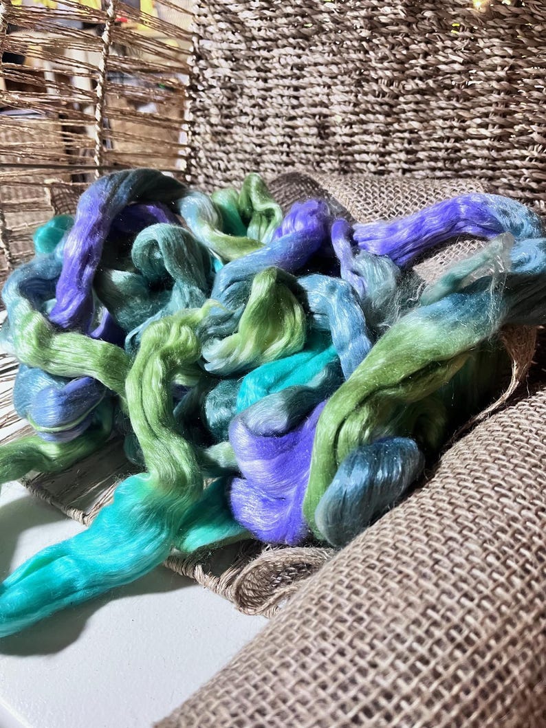 May include: A pile of colorful fiber in shades of teal, green, and purple. The fiber is arranged on a burlap-like fabric, with a woven basket in the background. The fiber appears soft and fluffy, suitable for crafting.