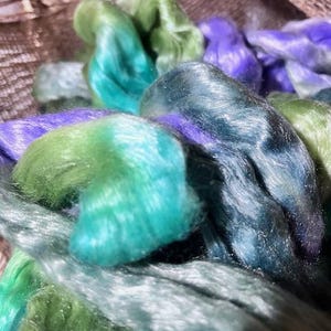 May include: Close-up of colorful silk fibers in shades of green, teal, and purple. The fibers are soft and shiny, with a slight sheen. The fibers are bundled together, creating a textured and layered effect. Ideal for fiber art projects.