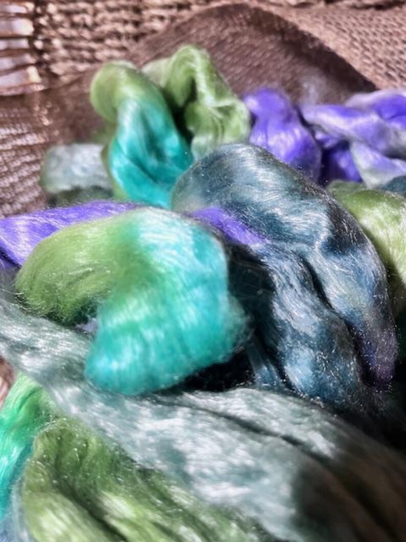 May include: Close-up of colorful silk fibers in shades of green, teal, and purple. The fibers are soft and shiny, with a slight sheen. The fibers are bundled together, creating a textured and layered effect. Ideal for fiber art projects.