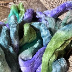 May include: Close-up of colorful fiber strands in shades of green, blue, and purple. The fiber is arranged in a basket, with a burlap background. The fibers appear soft and fluffy, ideal for crafting projects.