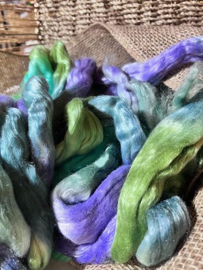 May include: Close-up of colorful fiber strands in shades of green, blue, and purple. The fiber is arranged in a basket, with a burlap background. The fibers appear soft and fluffy, ideal for crafting projects.