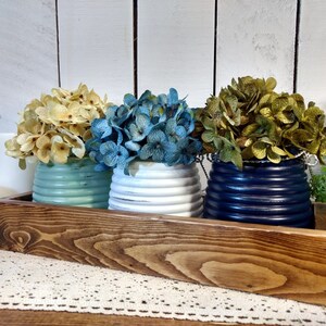 May include: Three decorative flower arrangements in different colored pots, sitting on a wooden tray. The flowers are yellow, blue, and green. The pots are painted in a distressed style.